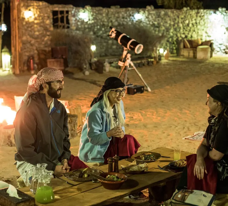 אורחים יושבים לארוחת ערב במדבר ליד טלסקופ